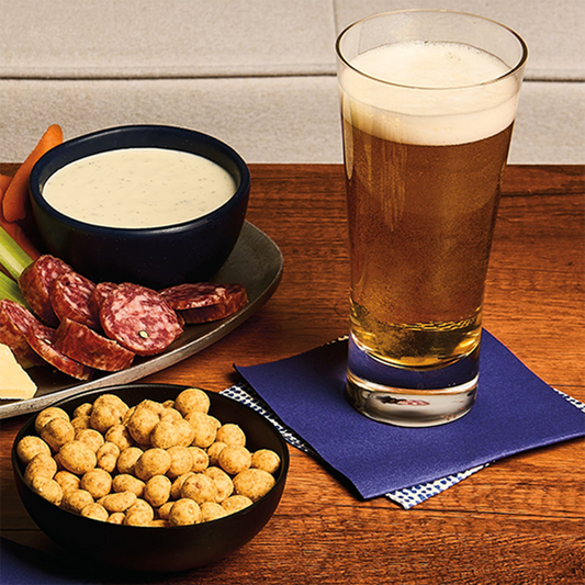 Glass of beer, bowl of snacks, and platter of charcuterie on a wooden table.